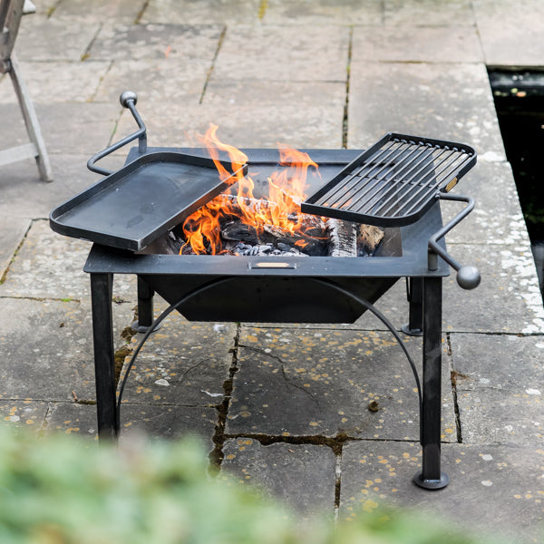 Firepits UK Box D square steel fire pit lit on a patio with both swing arm BBQ racks in place