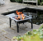 Box D square fire pit burning logs on a patio with the racks swung away