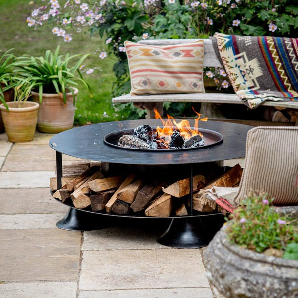Firepits UK Flat Ring of Logs 120 fire pit lit on a patio with logs stored around the base