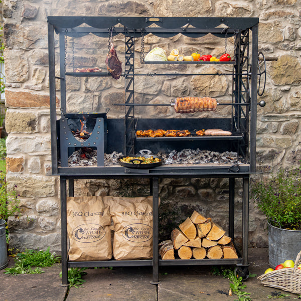 Firepits UK Asado Maximo fire table lit with food on the grill, rotisserie and hanging racks